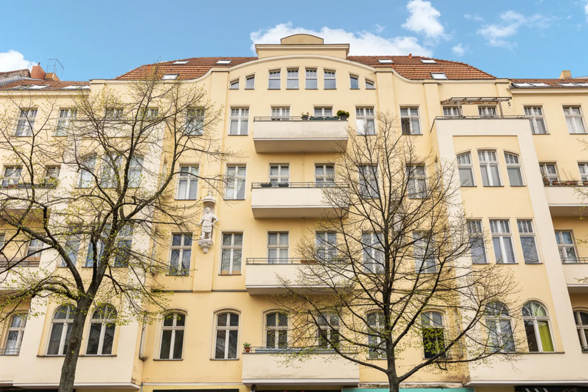 Mehrstöckiges Wohnhaus mit gelber Fassade, Balkonen und Bäumen davor.
