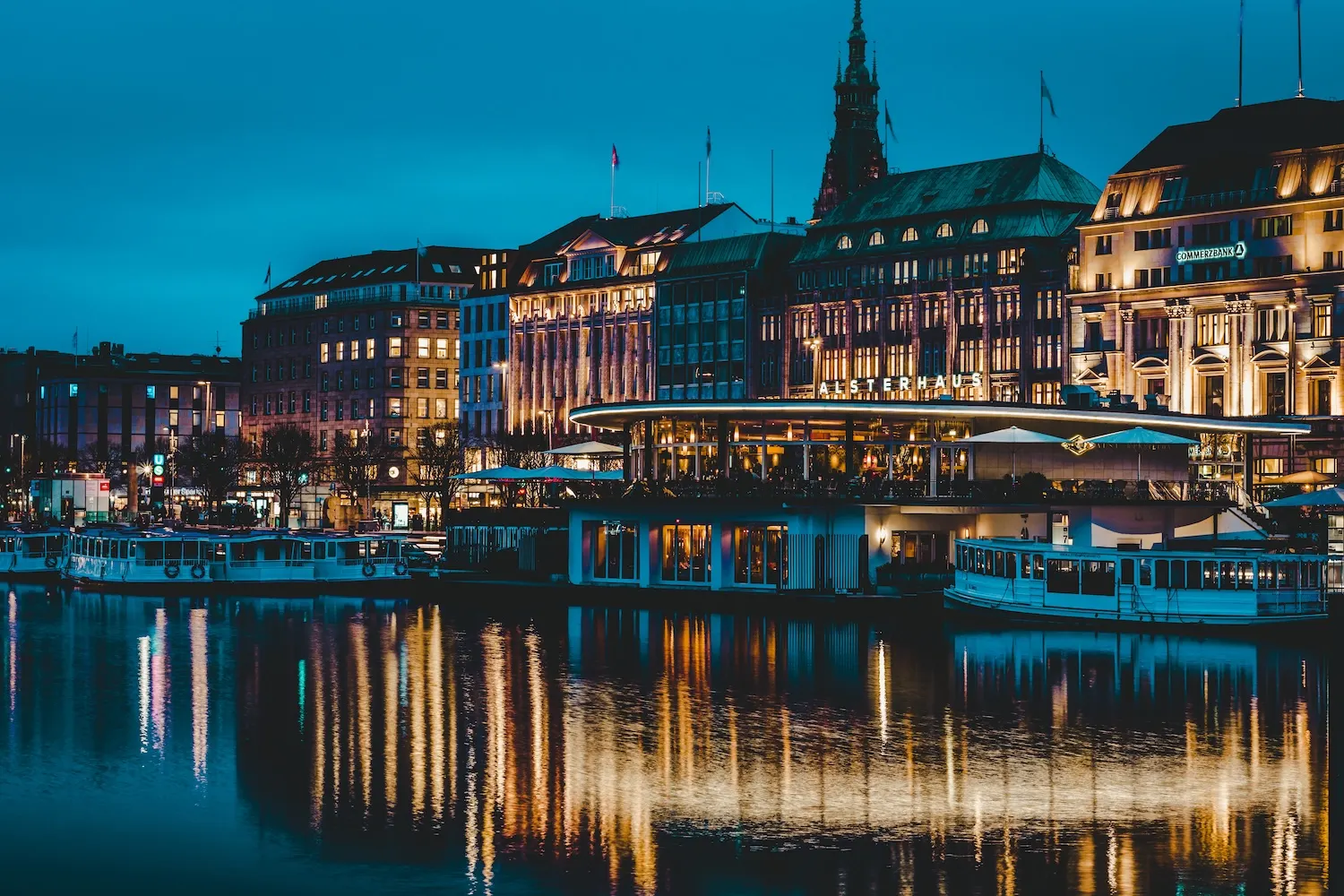 Beleuchtete Gebäude am Wasser bei Nacht mit Spiegelungen und Booten im Vordergrund.