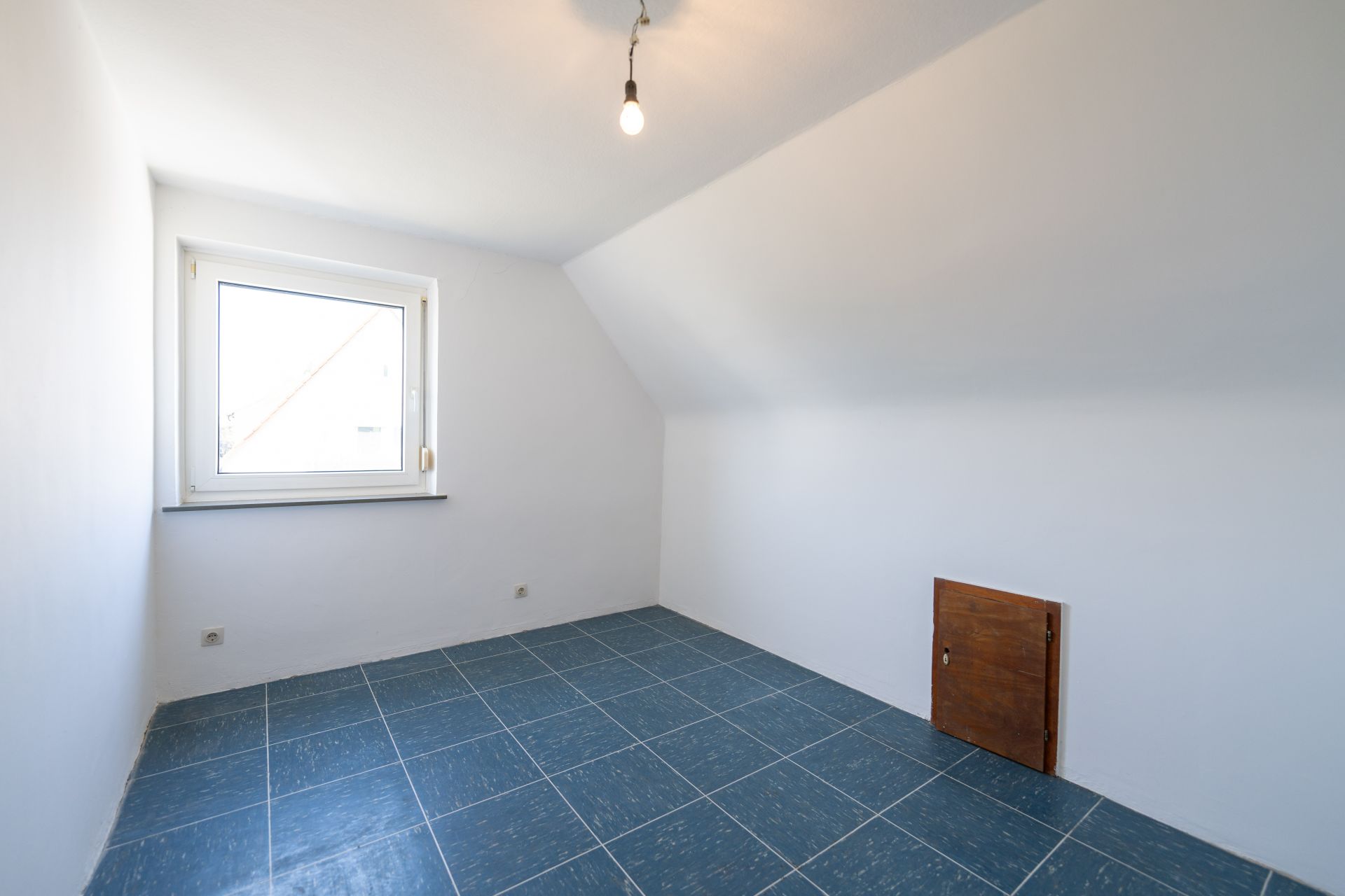 Leerer Raum mit blauen Fliesenboden, weißer Wand, Fenster und kleiner Holzklappe.