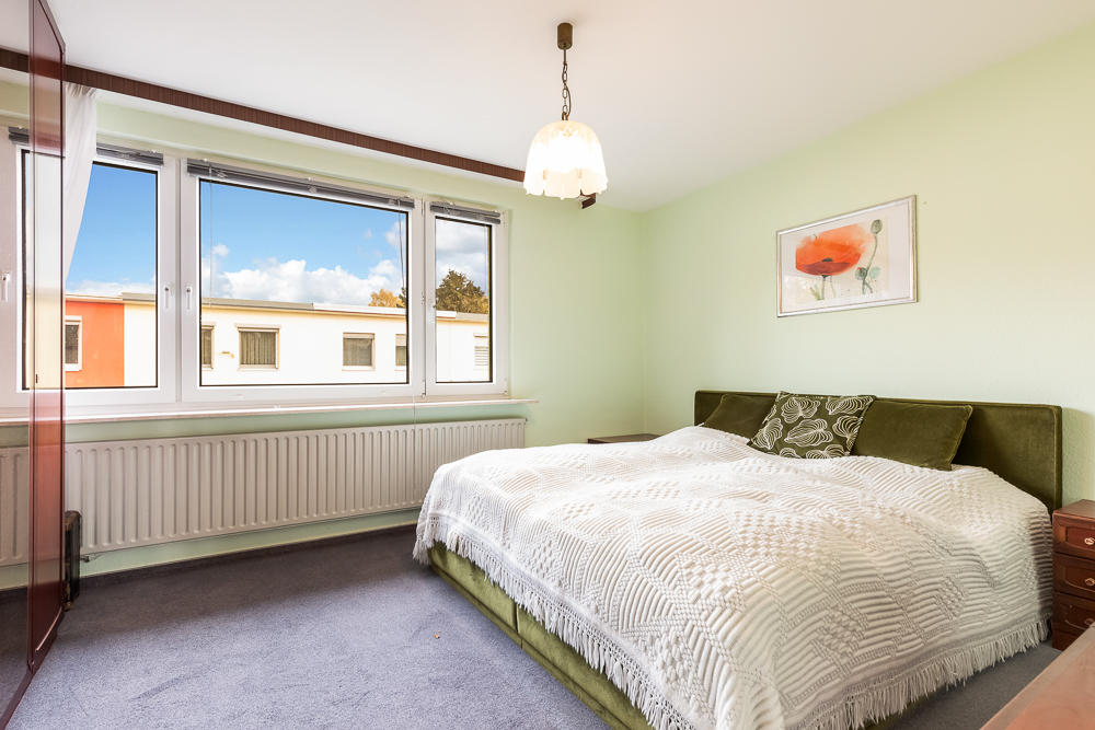 Helles Schlafzimmer mit großem Bett, drei Fenstern, grünem Kopfteil und Bild einer Blume an der Wand.