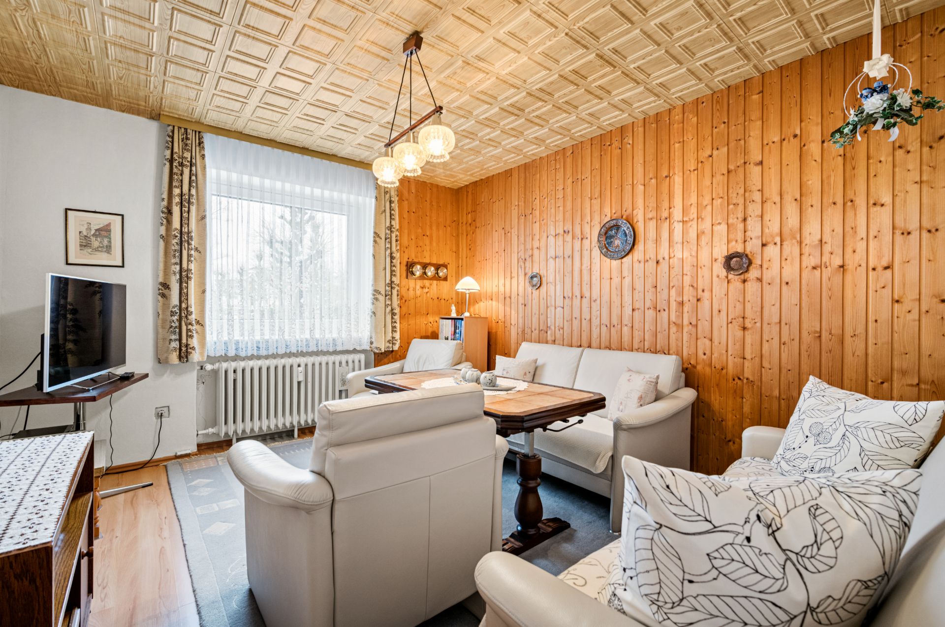 Wohnzimmer mit Holzvertäfelung, weißen Sofas, TV, Fenster und Deckenlampe.