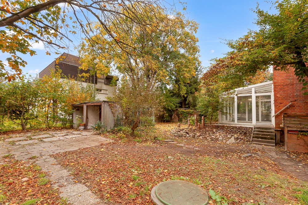 Garten mit Bäumen, Laub auf dem Boden, kleinem Schuppen und Wintergarten am Haus.