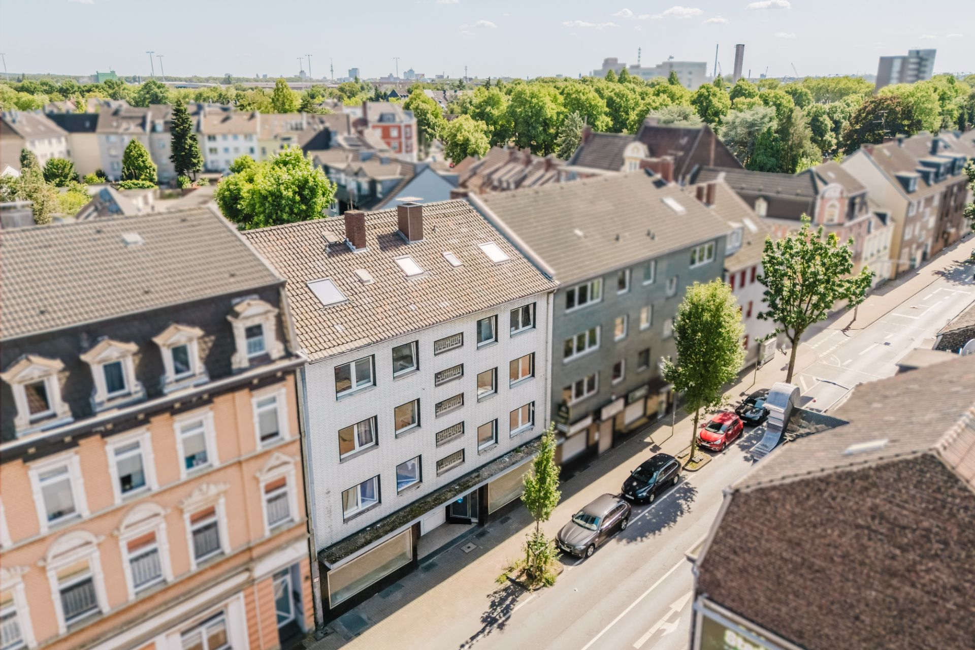 Straßenansicht mit Wohnhäusern, Bäumen und parkenden Autos an einer Hauptstraße.