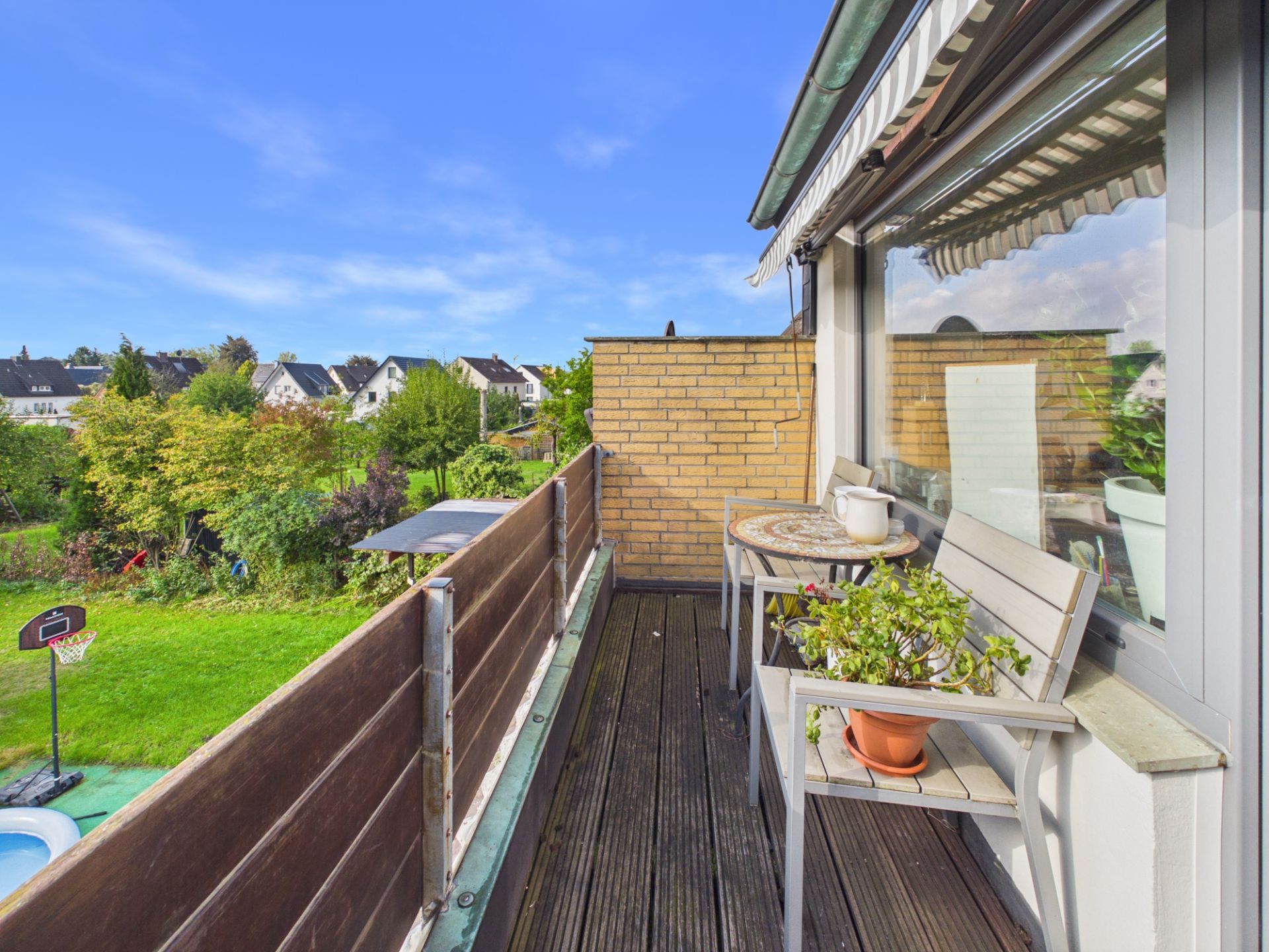 Ein Balkon mit Holzboden, Tisch und Bank, Pflanze im Topf, Blick auf Garten und Häuser.