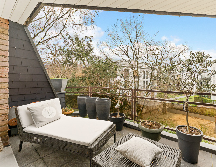 Balkon mit Liege, kleinem Tisch und mehreren Pflanzgefäßen, Blick auf Bäume und Gebäude.