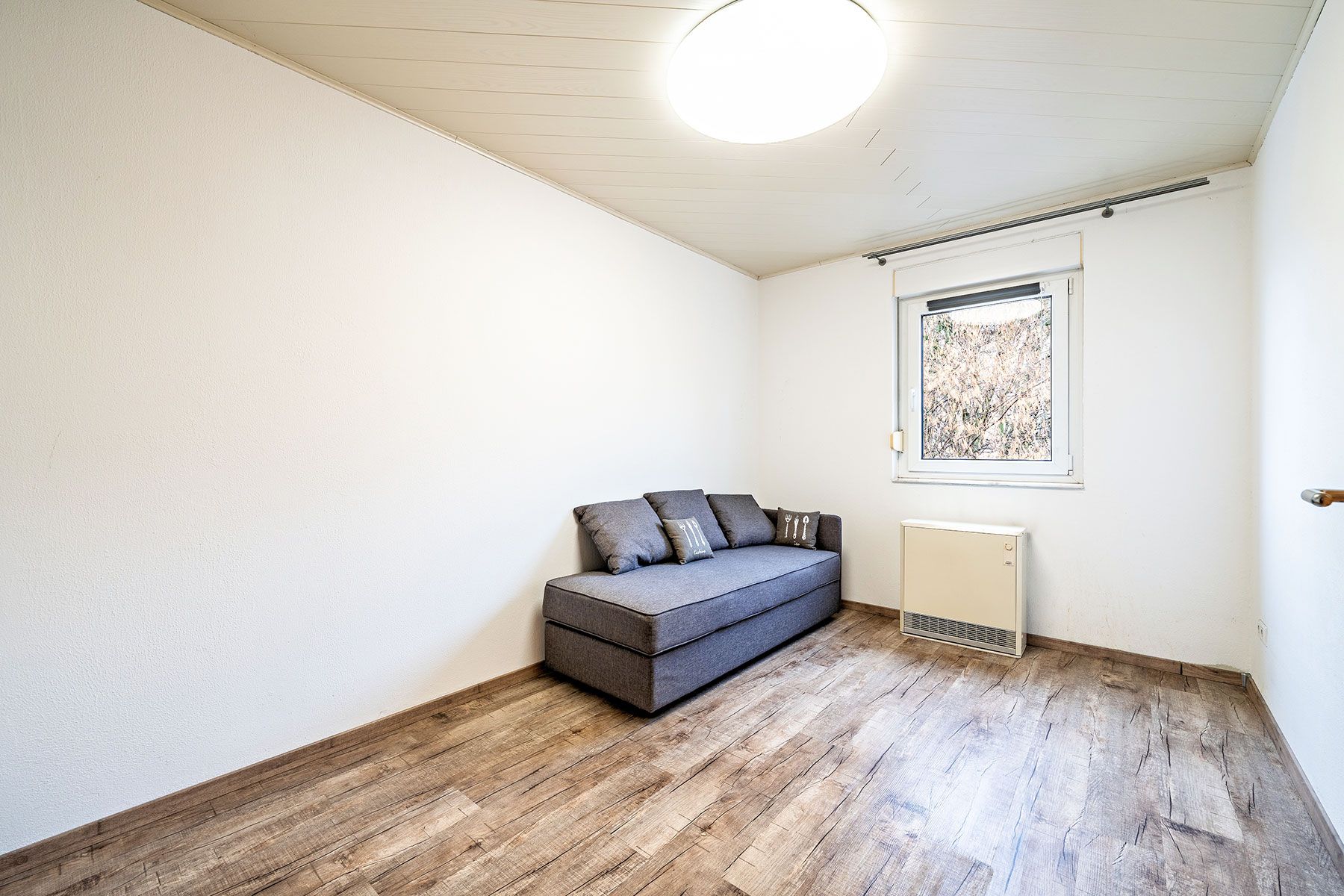 Helles Zimmer mit grauem Sofa, Holzfußboden und einem Fenster an der weißen Wand.