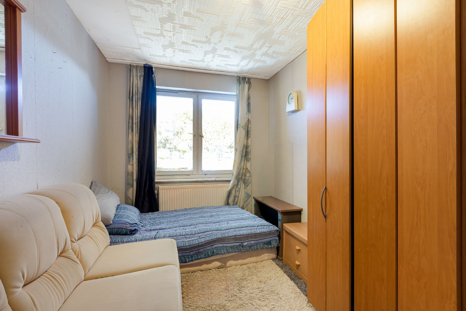 Kleines Schlafzimmer mit Bett, Sofa, Kleiderschrank und Fenster mit Vorhängen.
