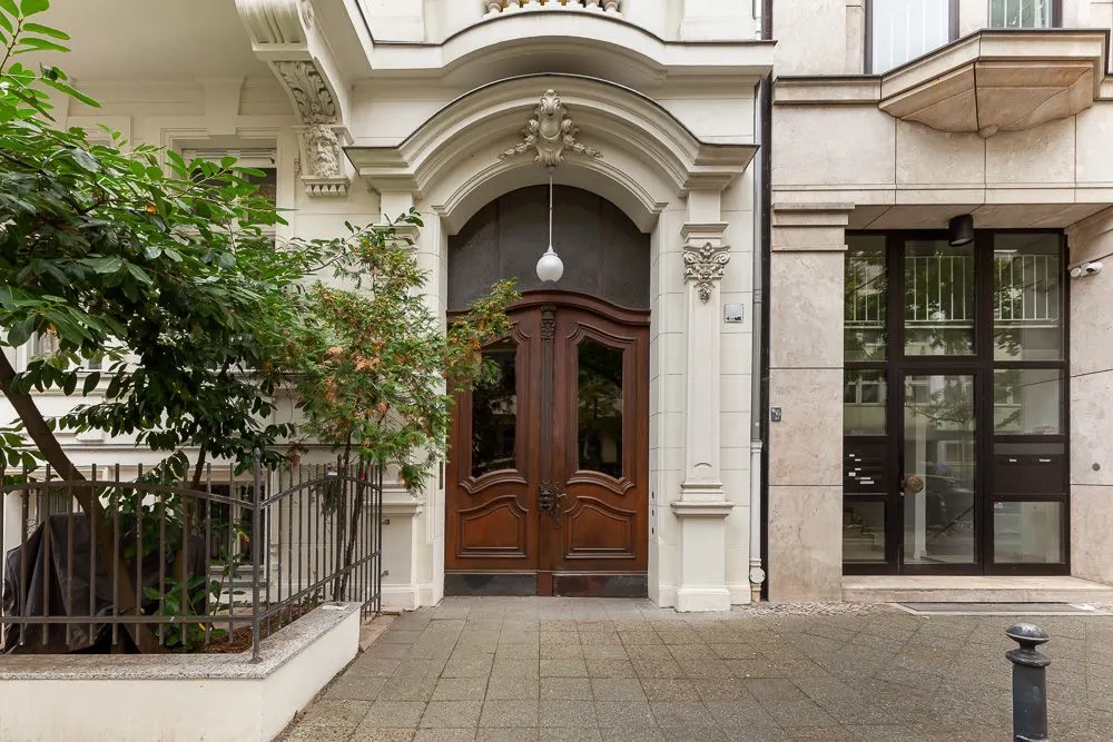 Große hölzerne Haustür mit Bogen, umgeben von verziertem weißen Mauerwerk und Pflanzen links.
