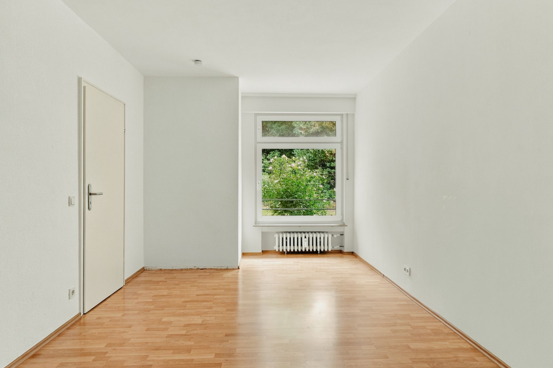 Leeres Zimmer mit Holzfußboden, weißer Wand, Tür links und großem Fenster mit Heizung darunter.