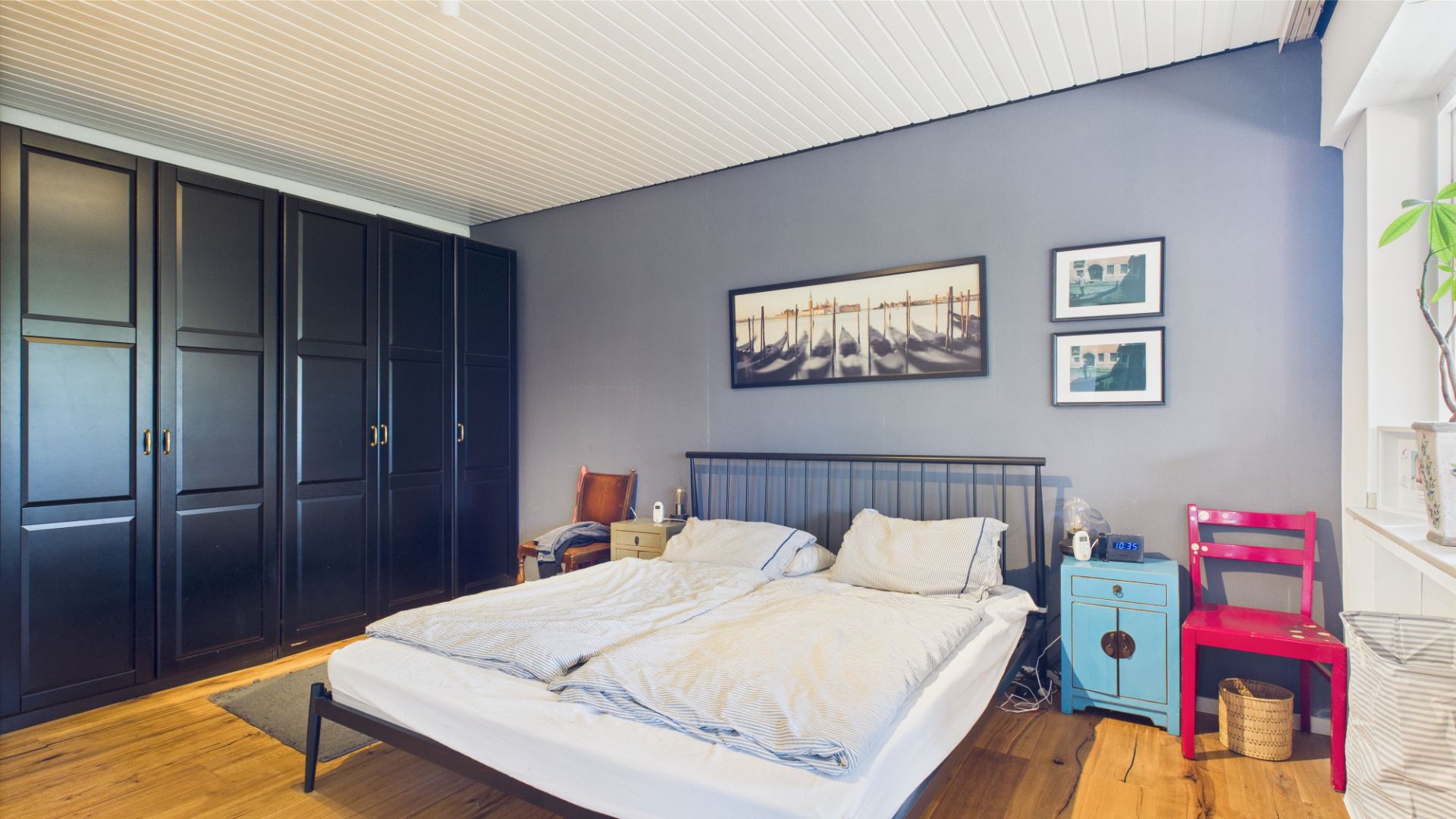 Schlafzimmer mit Doppelbett, schwarzem Kleiderschrank und bunten Nachttischen an einer blauen Wand.