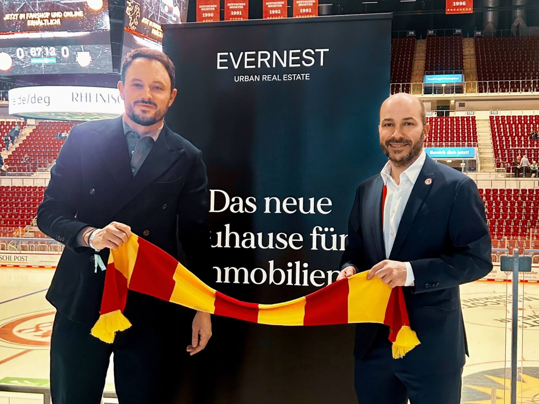 Zwei Männer halten einen rot-gelben Schal vor einem schwarzen Werbebanner in einer Eishalle.