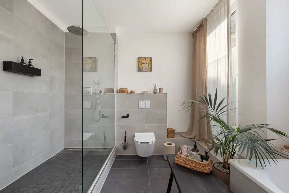 Modernes Badezimmer mit Dusche, WC, Pflanzen und großem Fenster mit Vorhang.