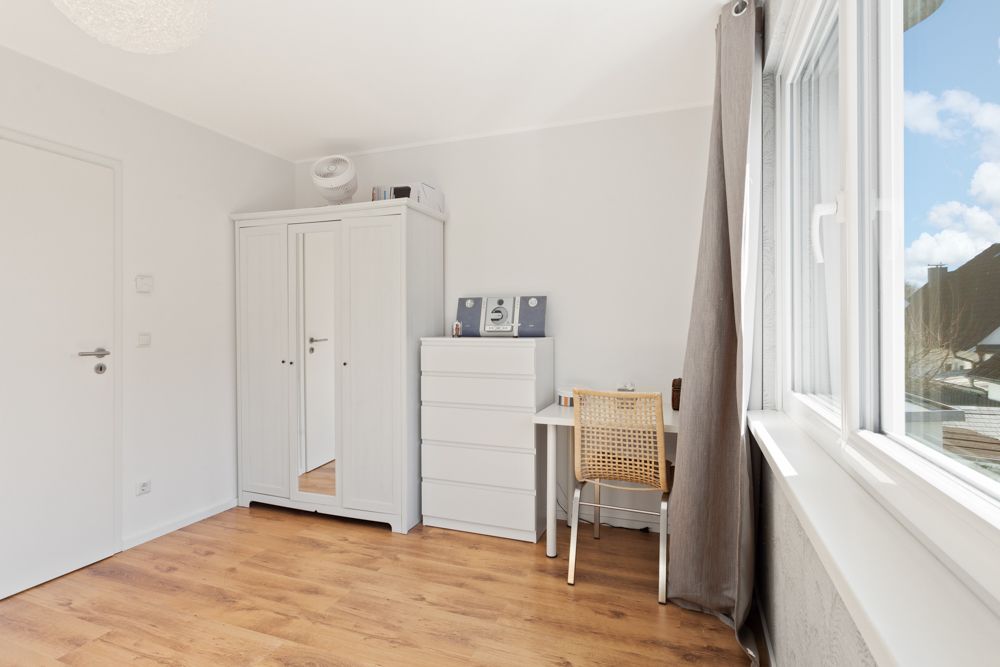 Helles Zimmer mit Holzfußboden, weißem Schrank, Kommode, Schreibtisch und Stuhl am Fenster.
