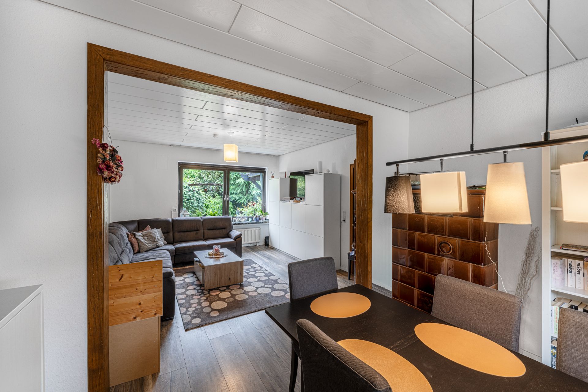 Wohnzimmer mit grauer Eckcouch, Teppich, Couchtisch und großem Fenster, angrenzender Essbereich mit Tisch und Stühlen.