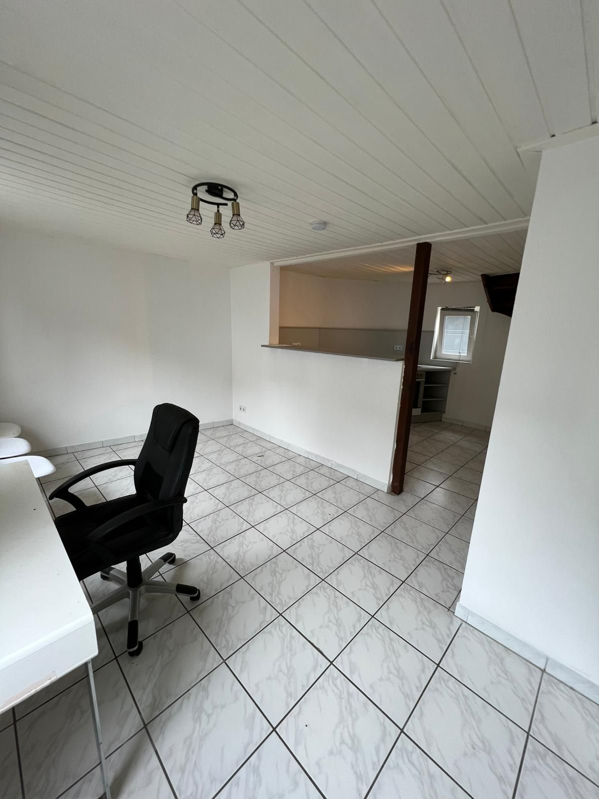 Heller Raum mit Fliesenboden, Schreibtisch und Bürostuhl, Durchgang zu weiterem Zimmer.