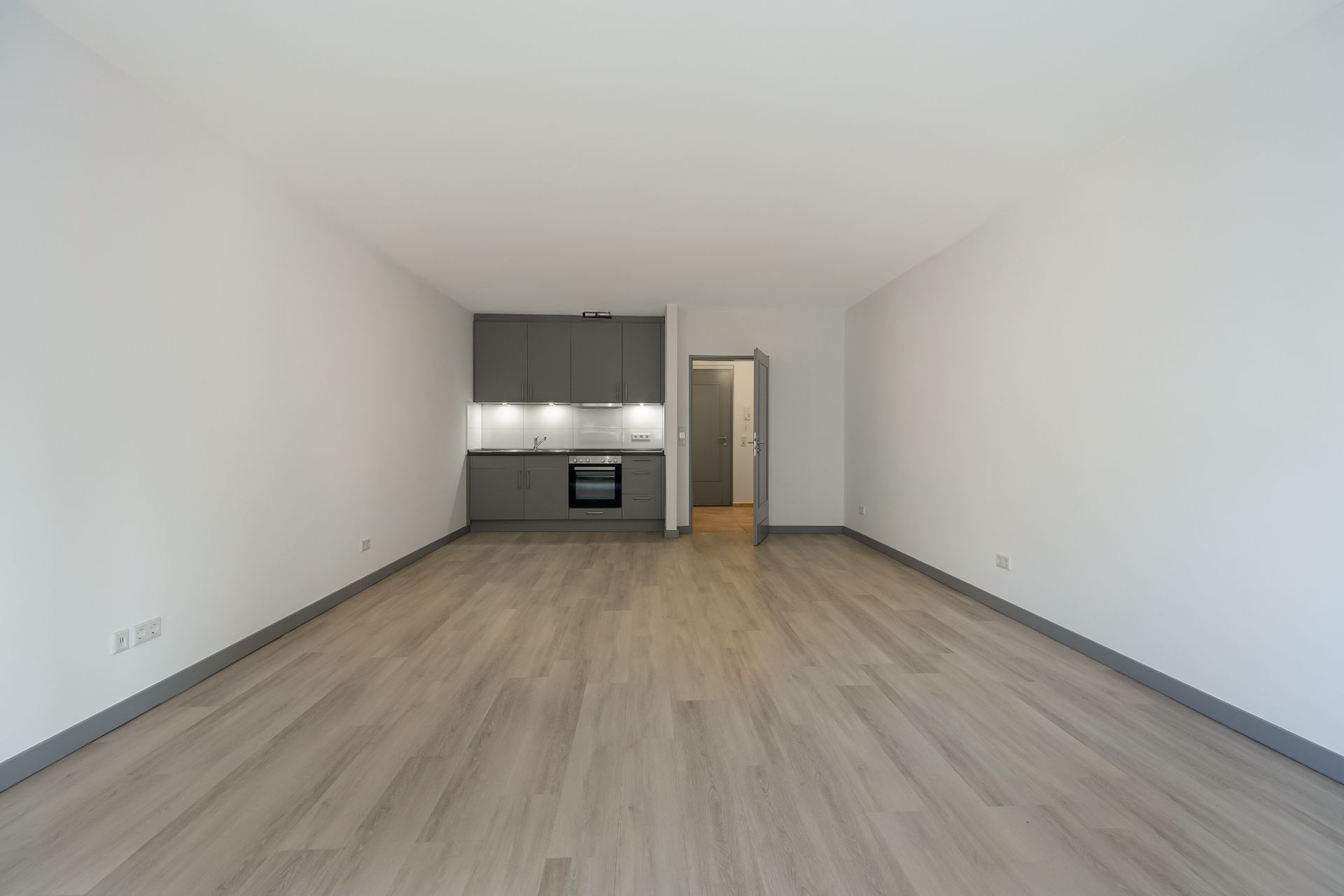 Leerer Raum mit Holzfußboden und offener grauer Einbauküche im Hintergrund.