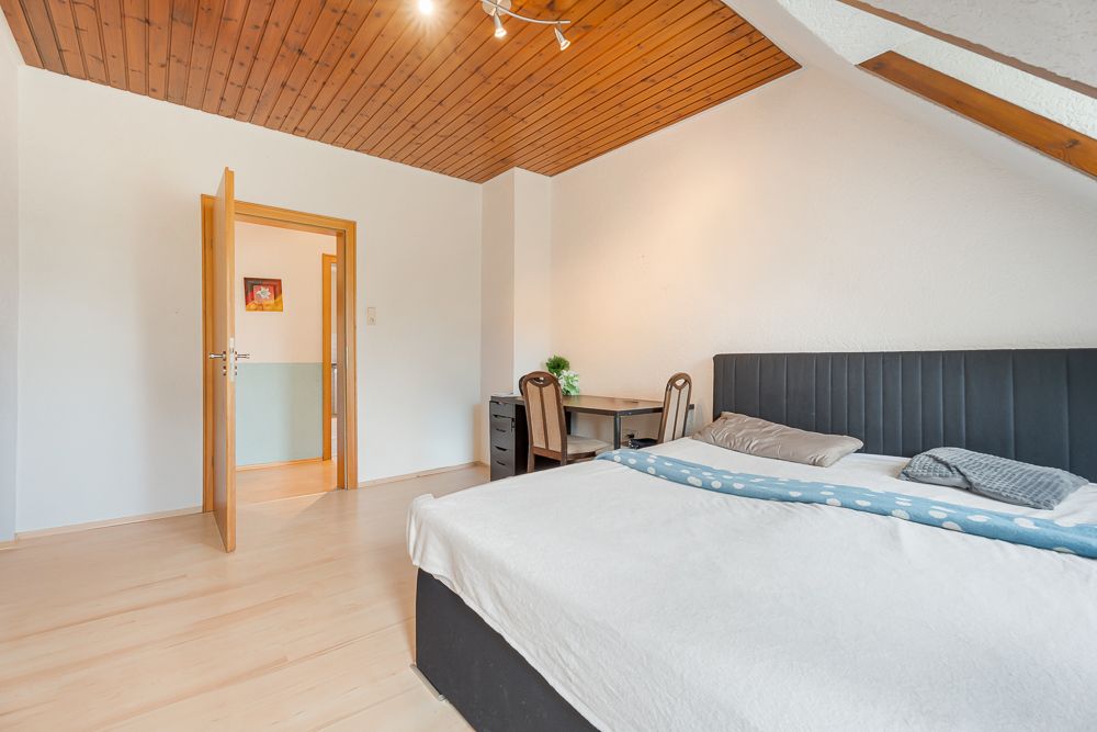 Schlafzimmer mit Doppelbett, Schreibtisch und offener Holztür, Holzdecke und helle Wände.