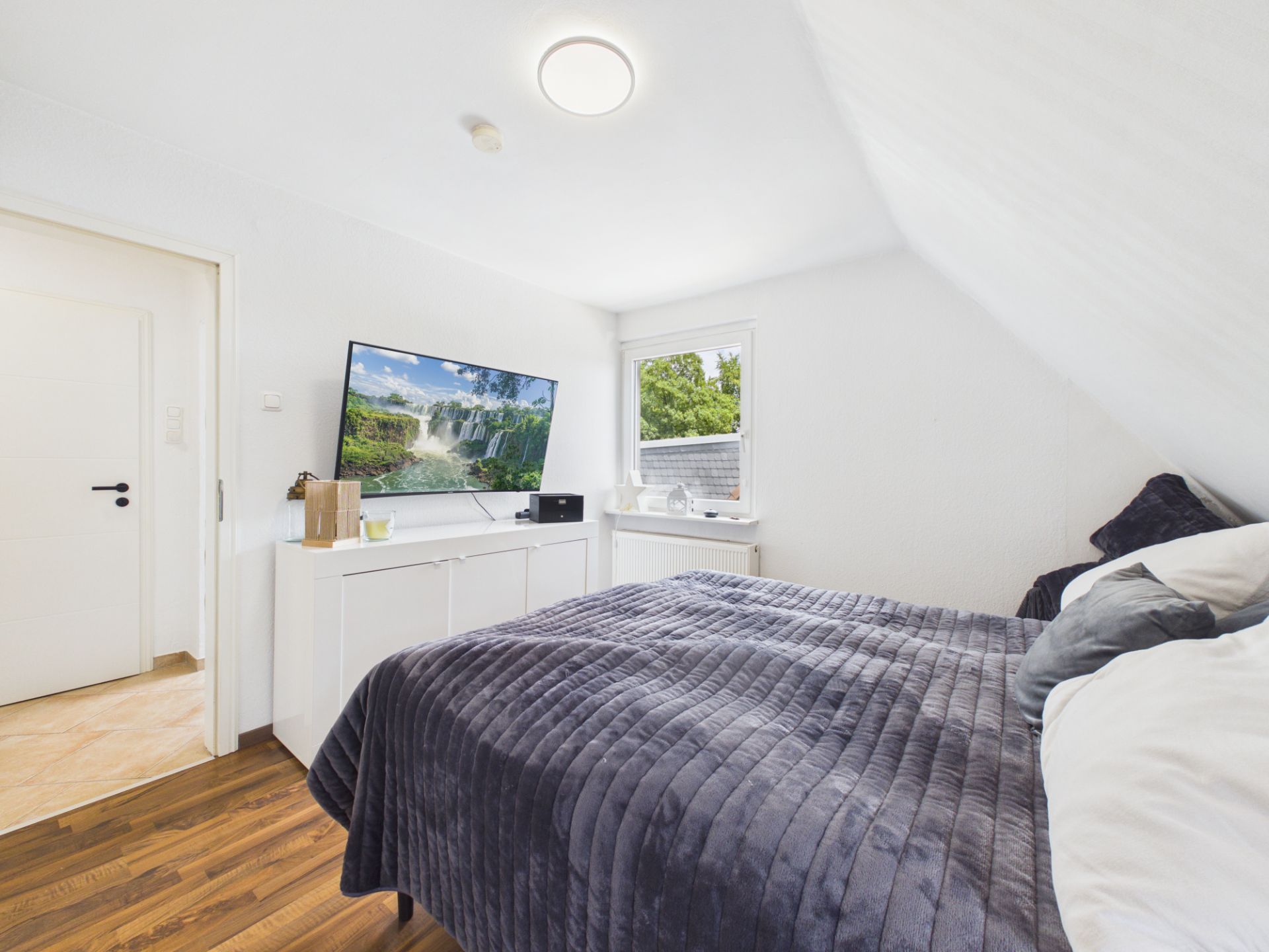 Helles Schlafzimmer mit großem Bett, TV auf einem weißen Sideboard und Fenster mit Blick ins Grüne.