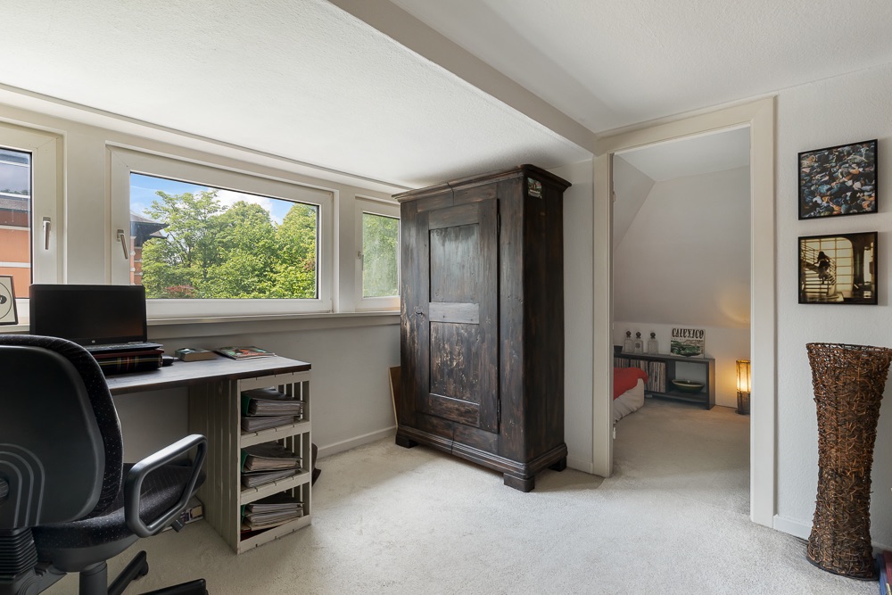 Helles Zimmer mit Schreibtisch, Stuhl, dunklem Schrank und Durchgang zu einem Schlafzimmer.