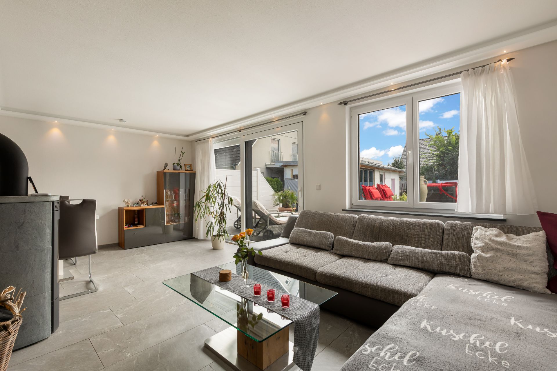 Helles Wohnzimmer mit großer Eckcouch, Glastisch, Fenster und Zugang zur Terrasse.