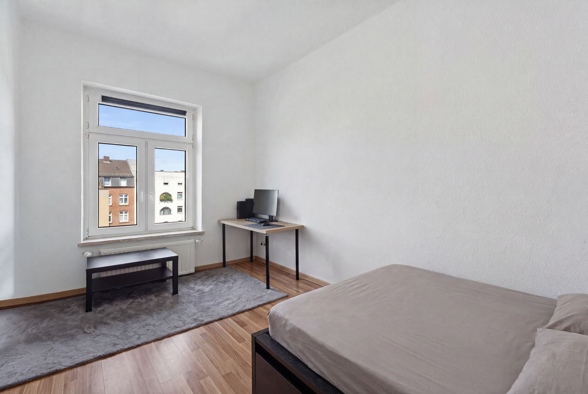 Helles Schlafzimmer mit Bett, kleinem Tisch, Schreibtisch und Computer am Fenster.