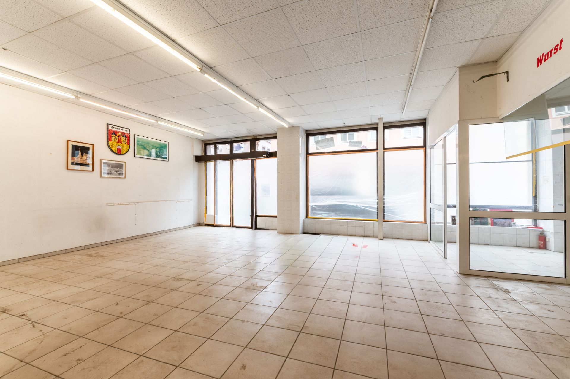 Leerer, gefliester Raum mit großen Fenstern und einigen Bildern an der Wand.