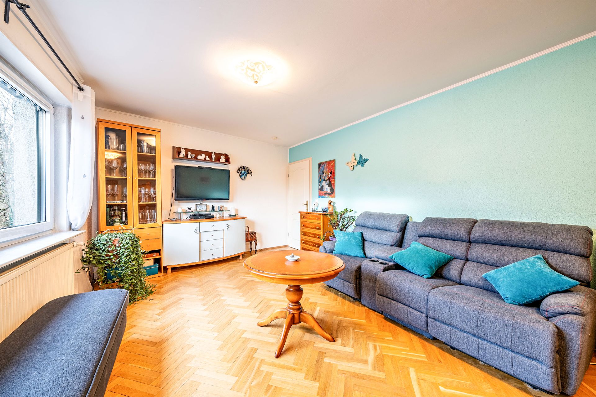 Helles Wohnzimmer mit grauem Sofa, Holztisch, Vitrine, Sideboard und großen Fenstern.