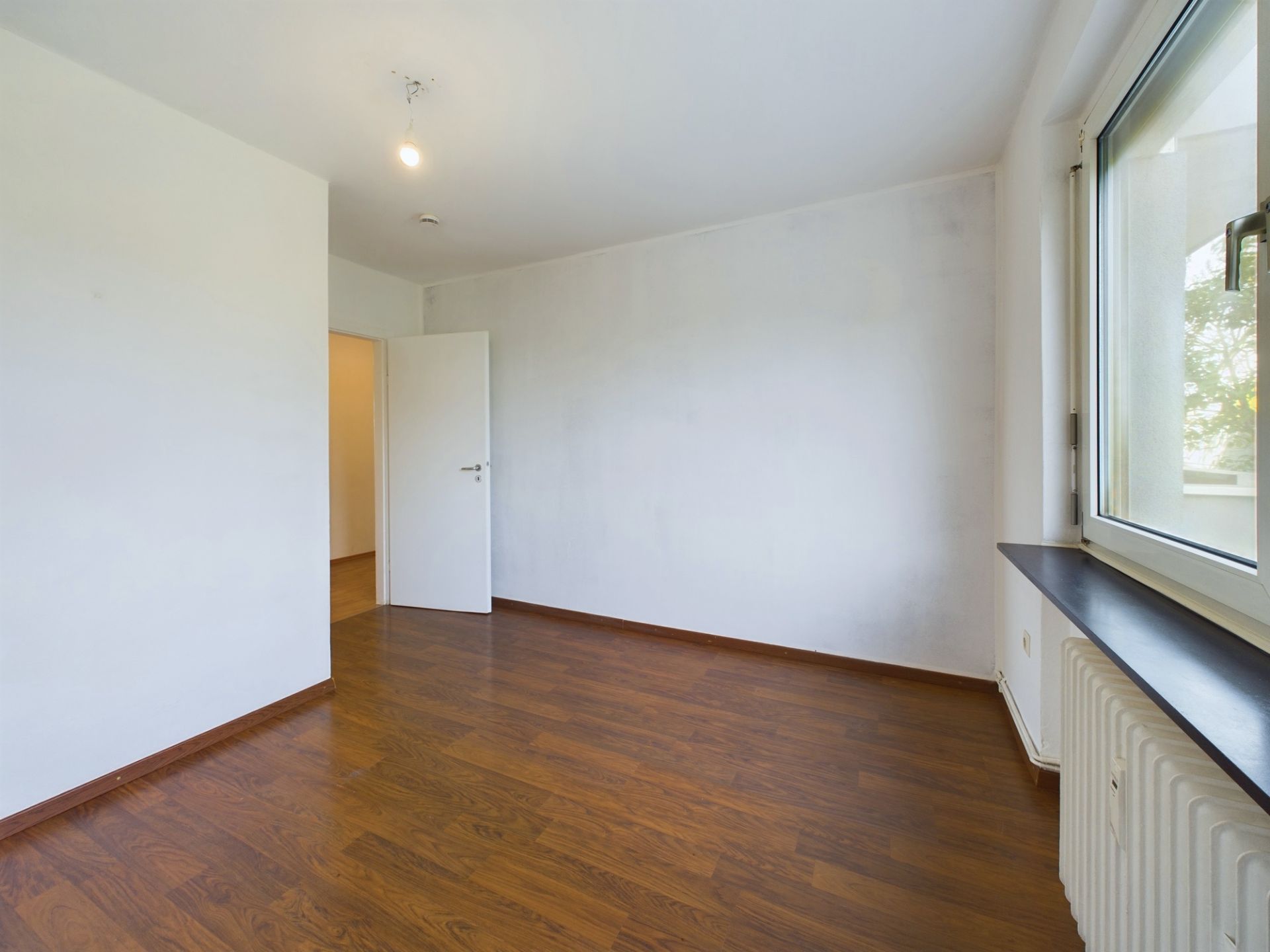 Leeres Zimmer mit weißer Wand, braunem Holzboden und großem Fenster.