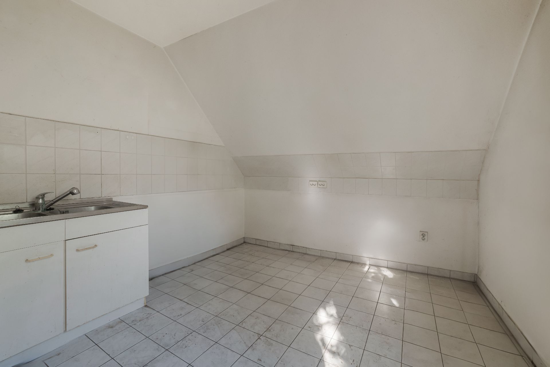 Leere Küche mit Fliesenboden, Spüle und weißem Unterschrank, schräge Decke.