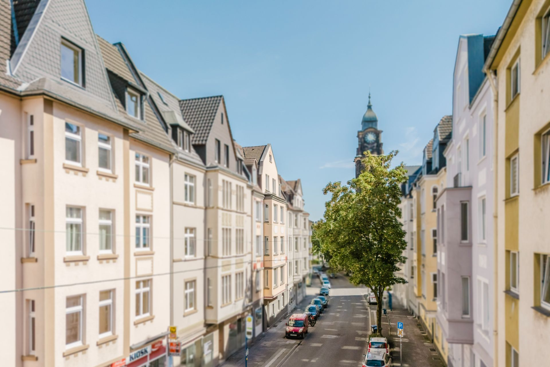 Blick auf eine ruhige Wohnstraße mit Bäumen, parkenden Autos und Mehrfamilienhäusern.