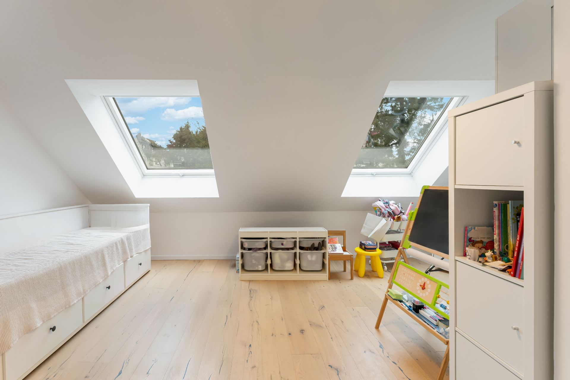 Helles Kinderzimmer mit Dachfenstern, Bett, Regal und Kindermöbeln auf Holzfußboden.