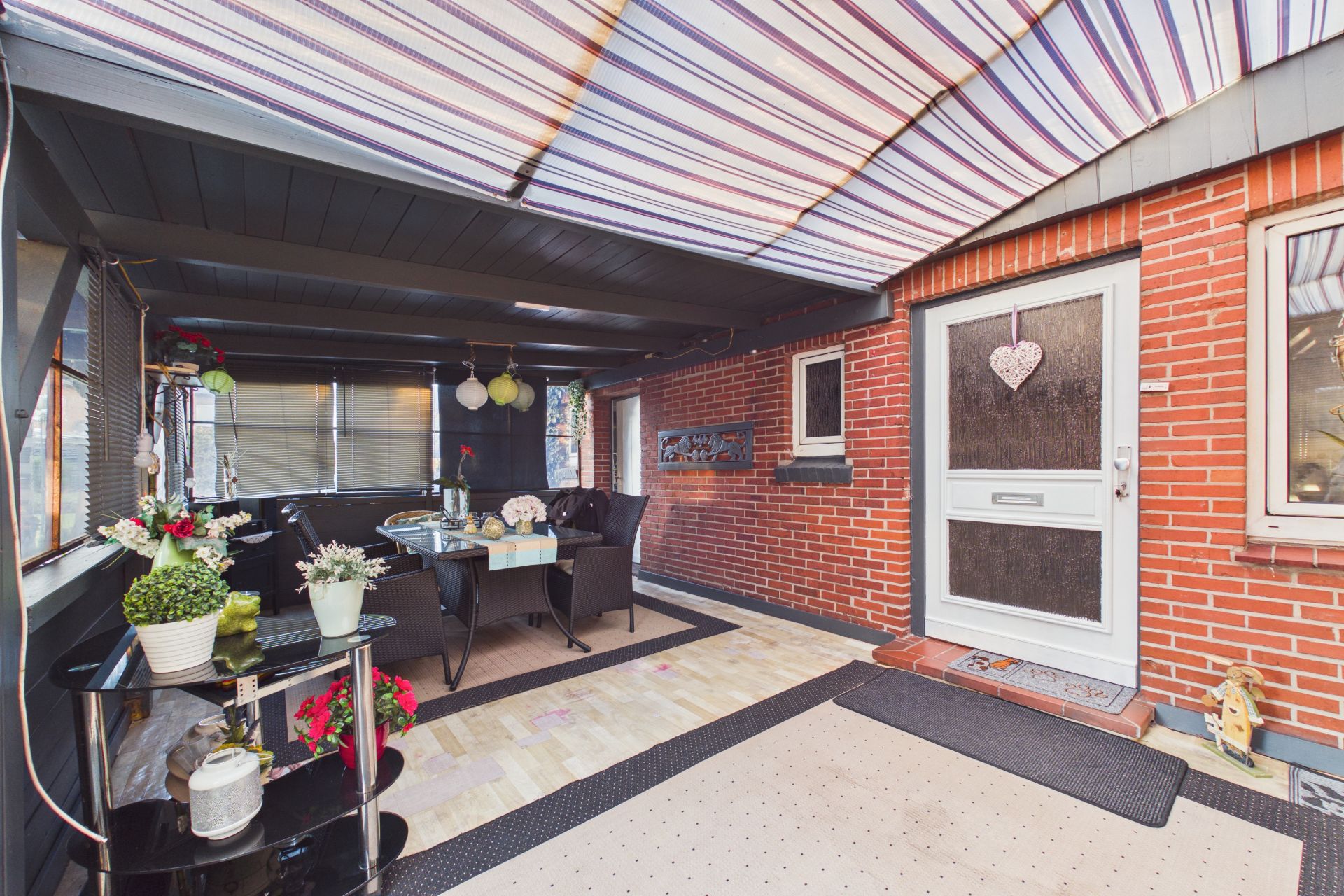 Überdachte Terrasse mit Esstisch, Stühlen, Pflanzen und Zugangstür zum Haus.