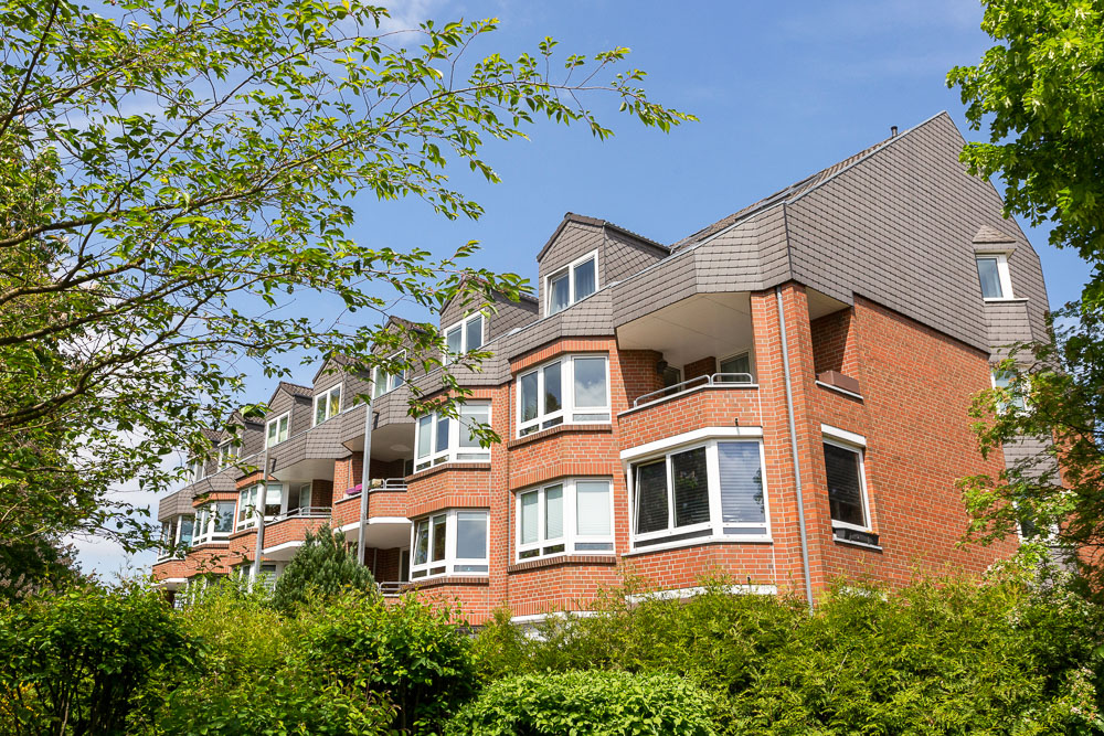 Mehrfamilienhaus aus rotem Backstein mit Balkonen, umgeben von Bäumen und Sträuchern.