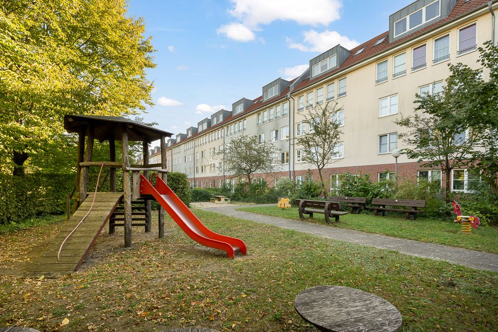 Spielplatz mit roter Rutsche und Holzklettergerüst neben Wohngebäude und Bänken.