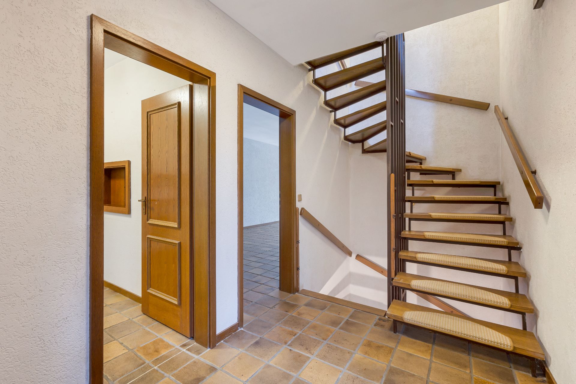Flur mit offener Holztreppe und zwei Türen, Fliesenboden, weiße Wände.