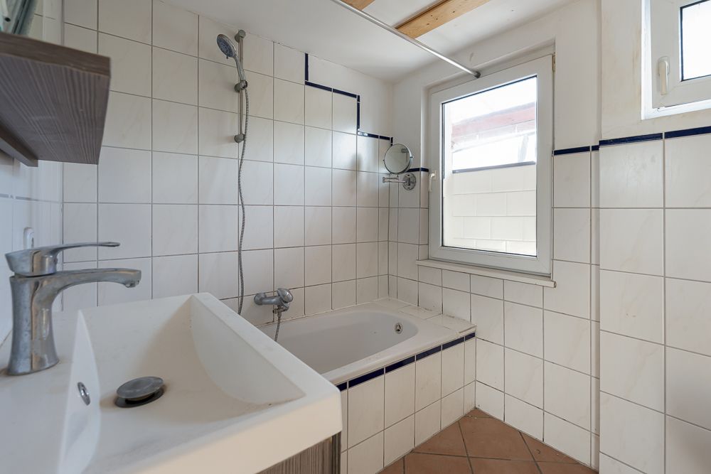Helles Badezimmer mit Badewanne, Waschbecken, zwei Fenstern und weißen Fliesen.