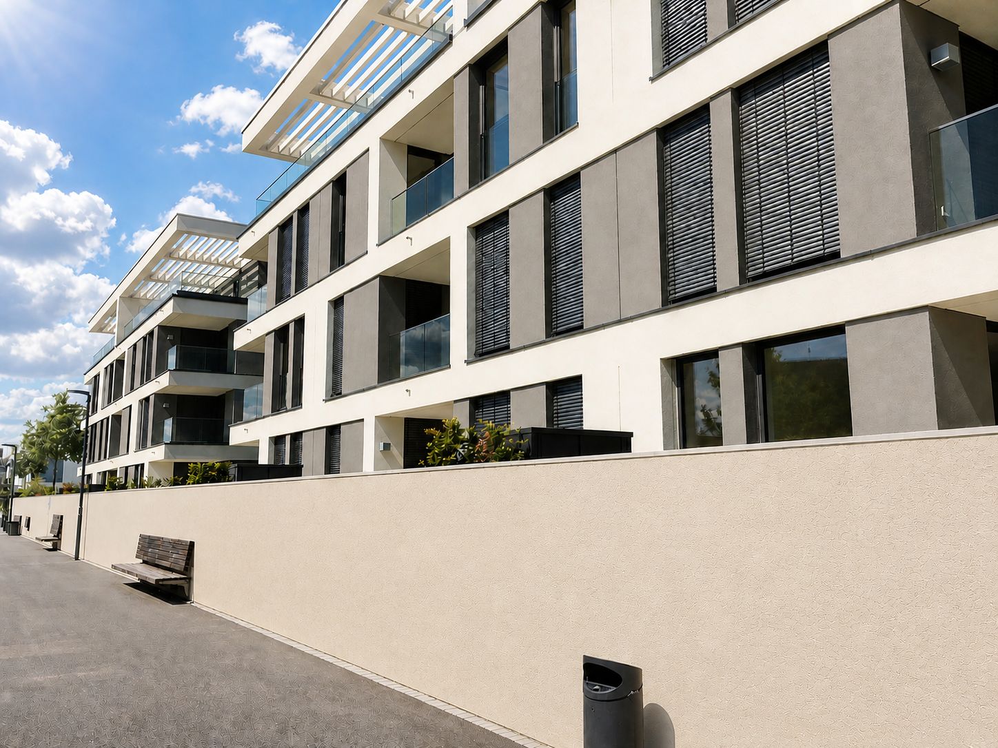 Modernes Wohngebäude mit Balkonen, Glasgeländern und Verschattungen, Gehweg mit Bank im Vordergrund.