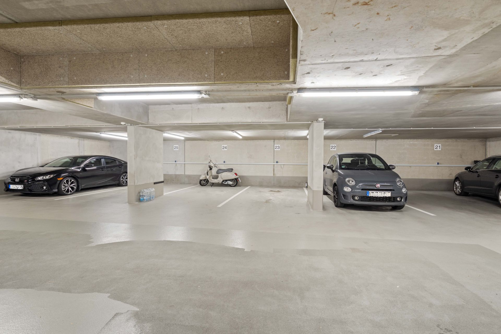 Tiefgaragenstellplätze mit Autos und einem Motorroller, helle Beleuchtung, Betonwände und -decke.