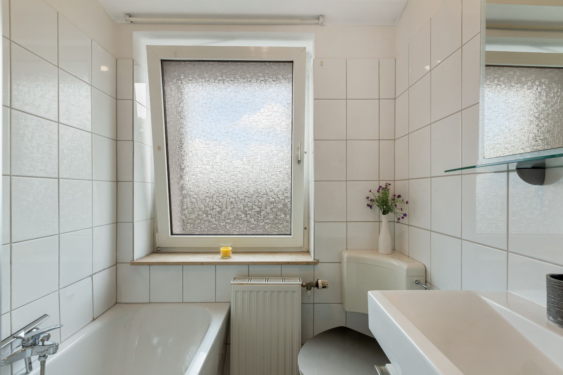 Kleines Badezimmer mit Badewanne, Fenster, Waschbecken und WC, weiß gefliest.