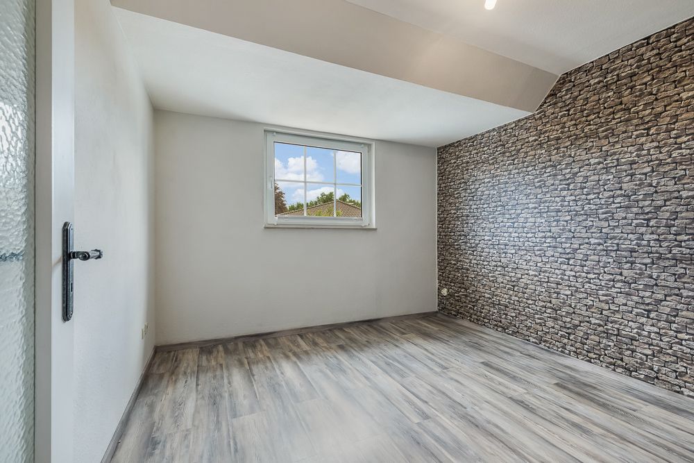 Leeres Zimmer mit Holzfußboden, Fenster und einer Wand in Steinoptik.