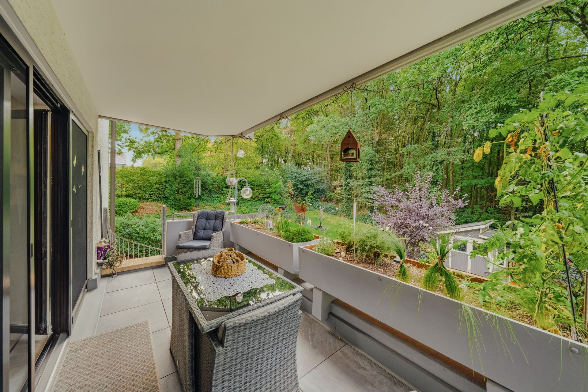 Überdachte Terrasse mit Sitzgruppe, Tisch und Blick auf einen grünen Garten mit Bäumen.