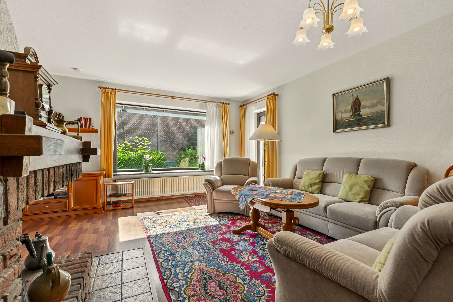 Helles Wohnzimmer mit Teppich, Sofa, Sesseln, Kamin und großem Fenster mit Blick auf den Garten.