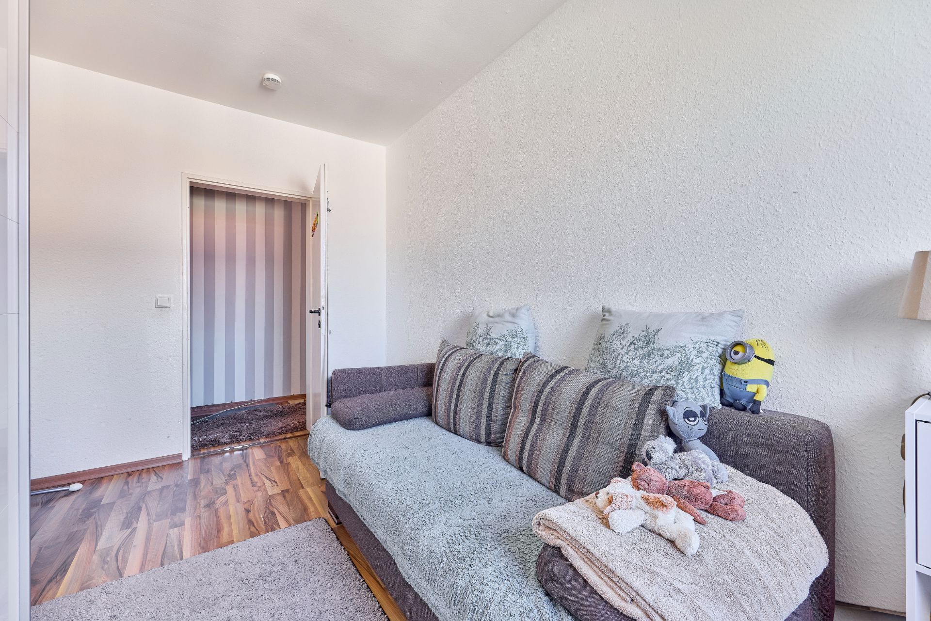 Kleines Zimmer mit grauem Sofa, Kuscheltieren und Holzfußboden.