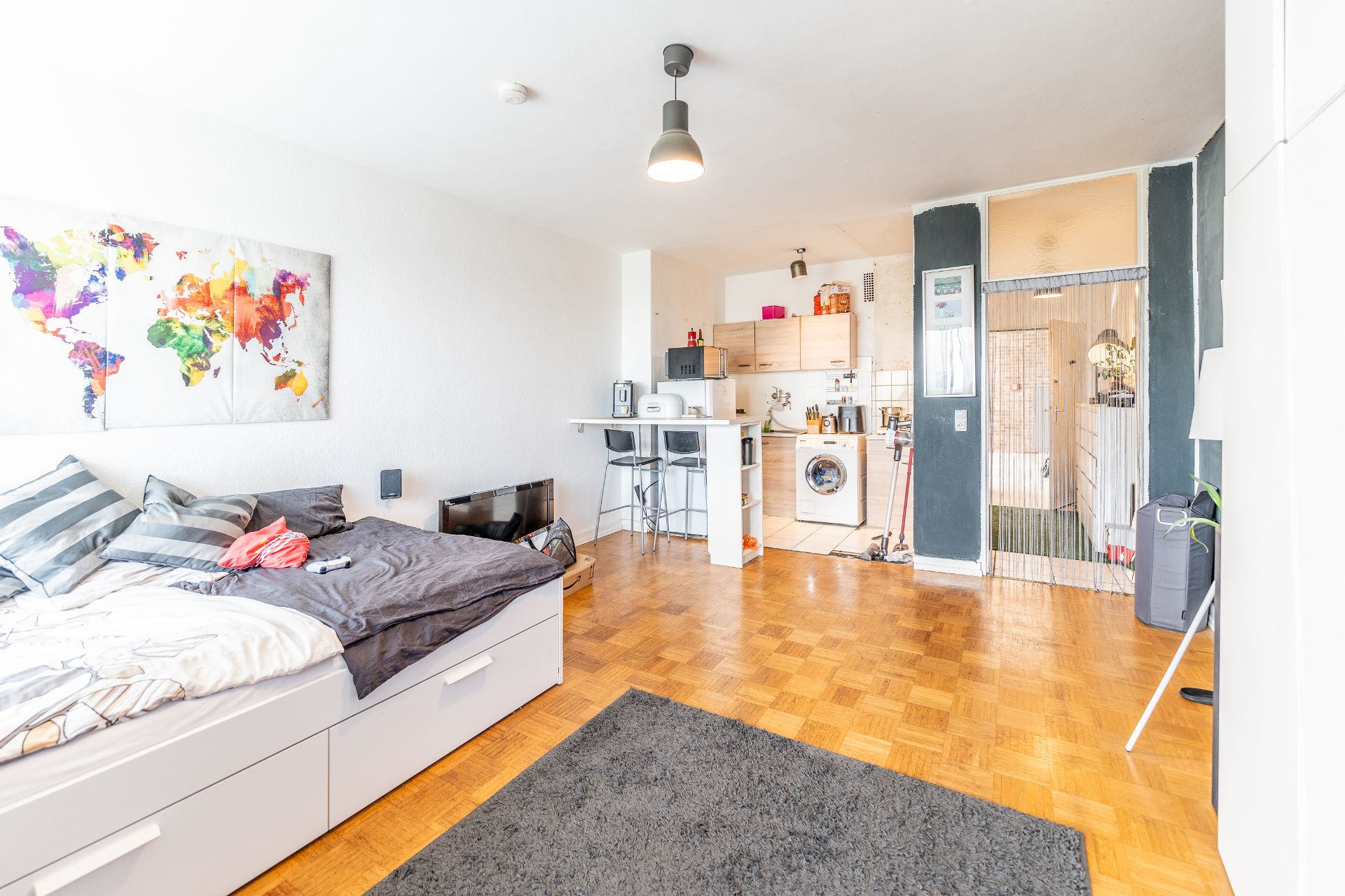 Helles Zimmer mit Bett, Küchenzeile, Barhockern und einem Fernseher auf Parkettboden.