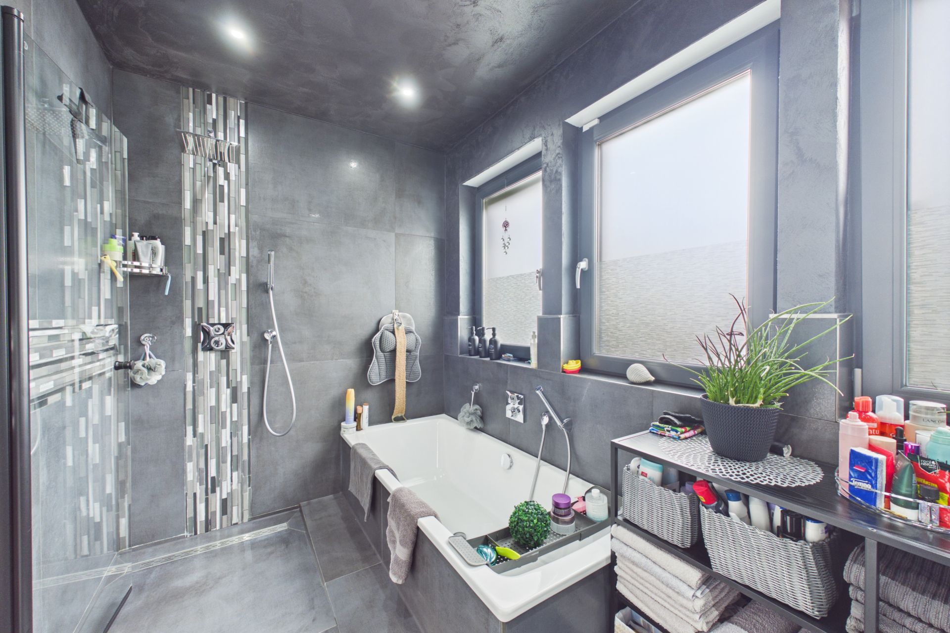 Modernes Badezimmer mit Badewanne, Dusche, zwei Fenstern und Regal mit Pflegeprodukten.