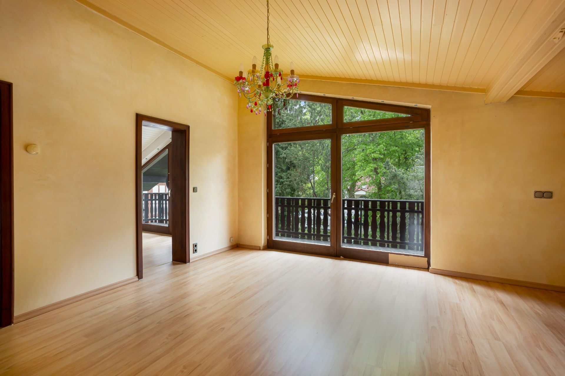 Leerer Raum mit Holzfußboden, Kronleuchter und großen Fenstern mit Blick auf einen Balkon und Bäume.