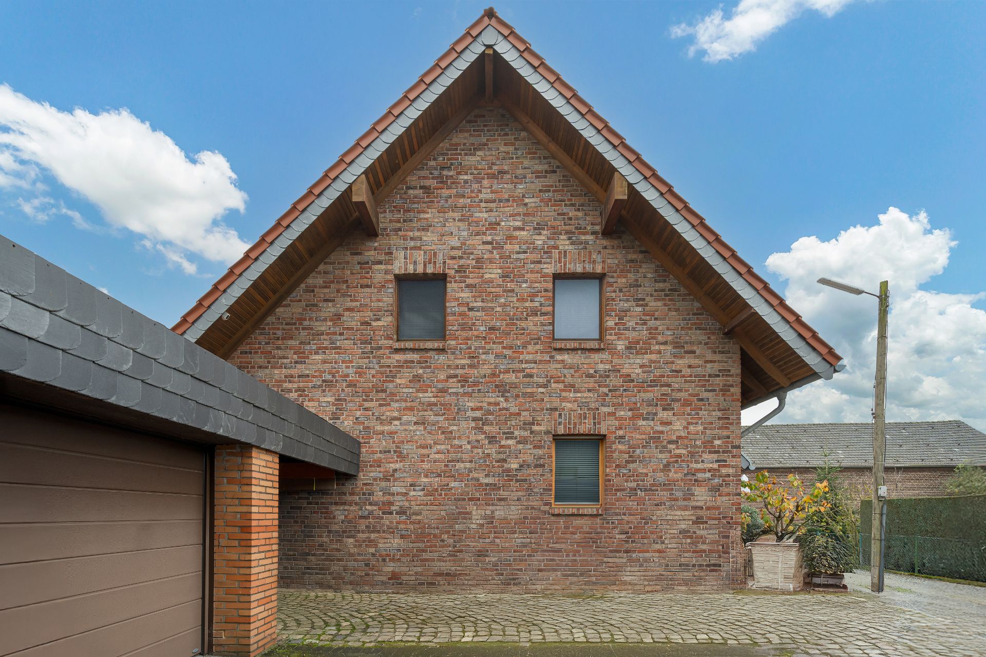 Backsteinfassade eines Wohnhauses mit Satteldach und drei Fenstern, davor gepflasterter Hof.