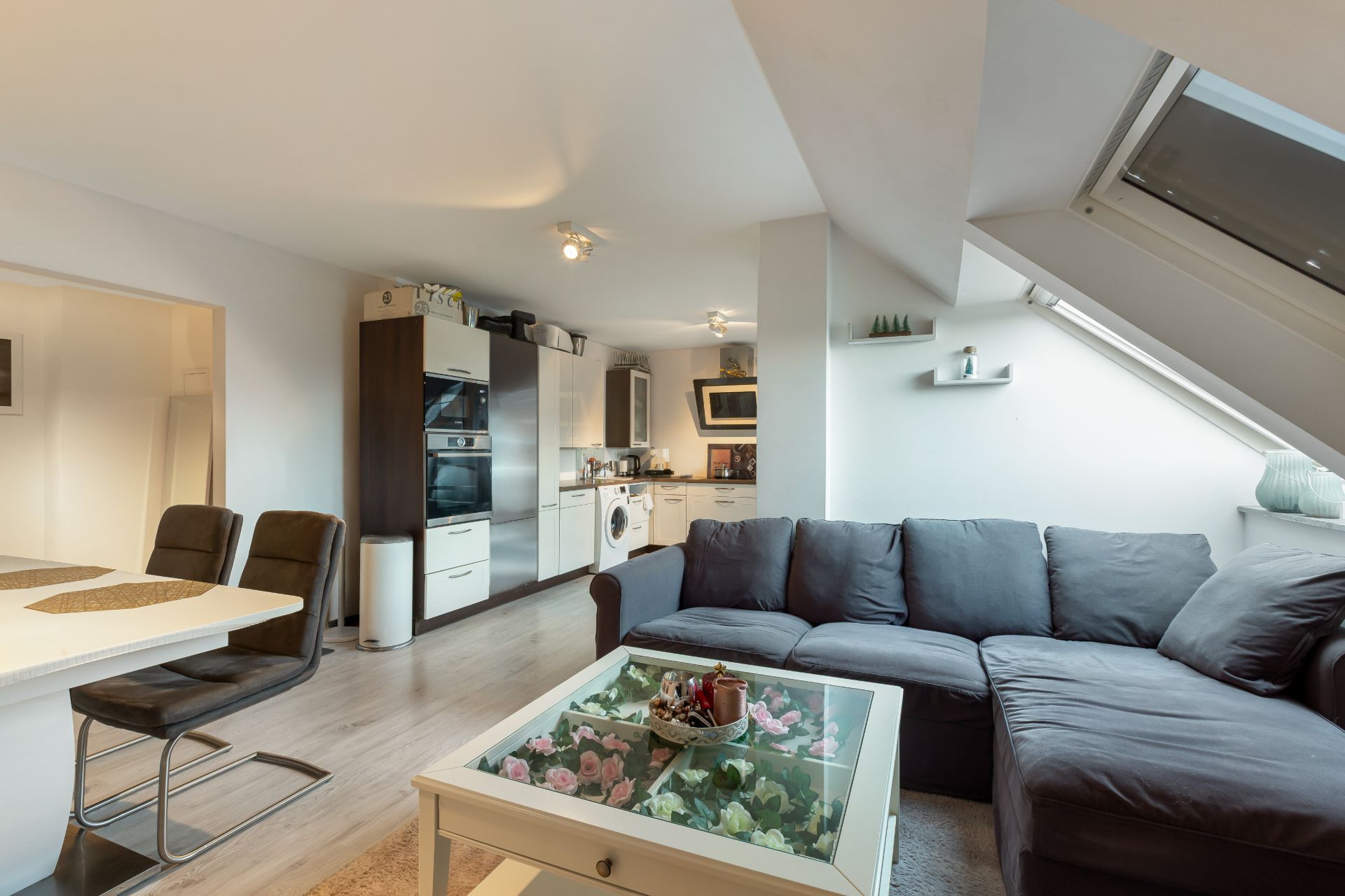Modernes Wohnzimmer mit Ecksofa, Glastisch und offener Küche unter Dachschräge.