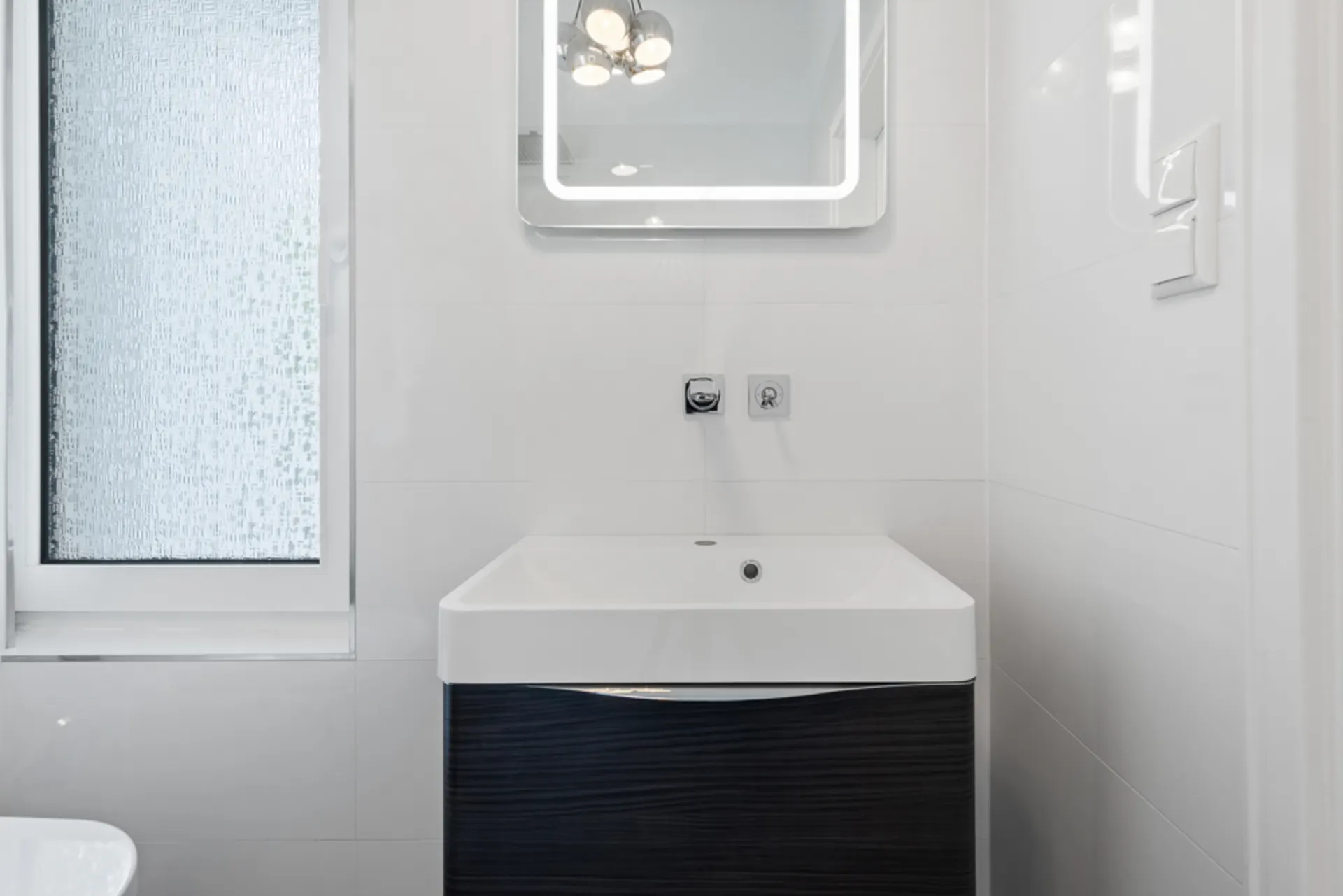 Modernes Badezimmer mit Waschbecken, beleuchtetem Spiegel und Fenster mit Milchglas.