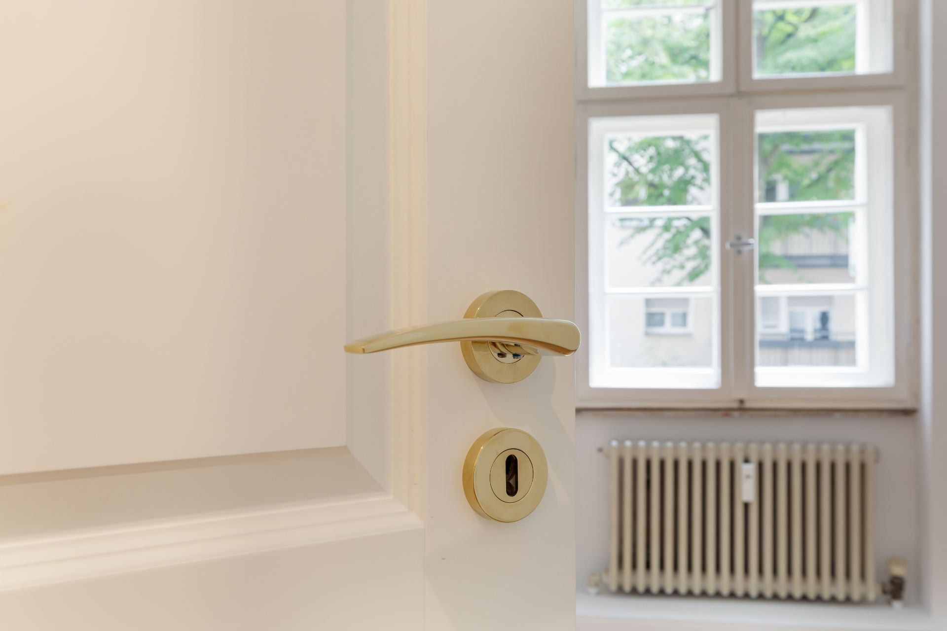 Weiße Tür mit goldenem Griff vor Fenster und Heizkörper in einem Raum.